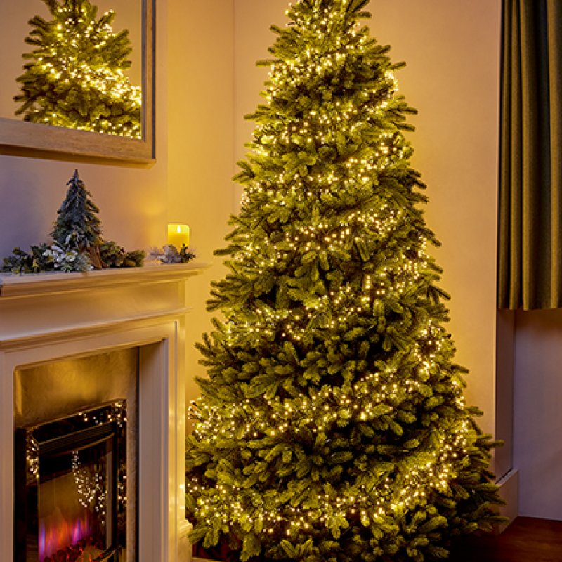 A tall artificial Christmas tree decorated with warm white string lights, standing beside a lit fireplace in a cozy indoor setting.