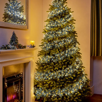 A tall artificial Christmas tree with white  and warm white string lights, styled in a cosy living room next to a fireplace with a mirror showing its reflection.