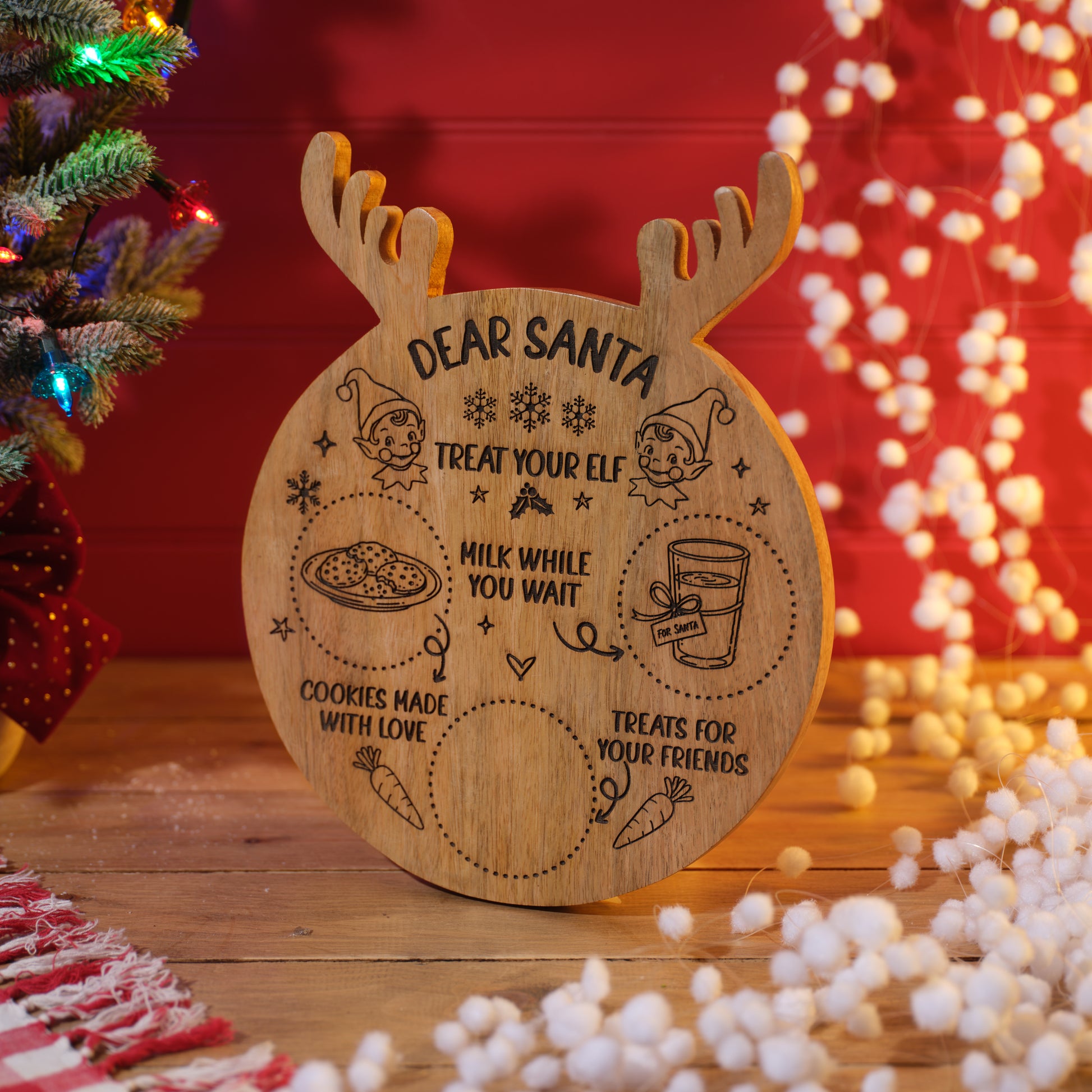 A round wooden Christmas treat mango wood board with antlers, engraved with 'DEAR SANTA TREAT YOUR ELF' and holiday illustrations, displayed on a wooden surface with a blurred Christmas tree and warm lights in the background.