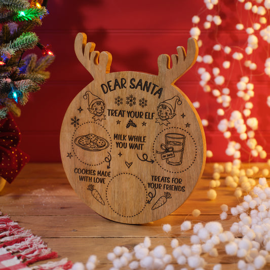 A round wooden Christmas treat mango wood board with antlers, engraved with 'DEAR SANTA TREAT YOUR ELF' and holiday illustrations, displayed on a wooden surface with a blurred Christmas tree and warm lights in the background.