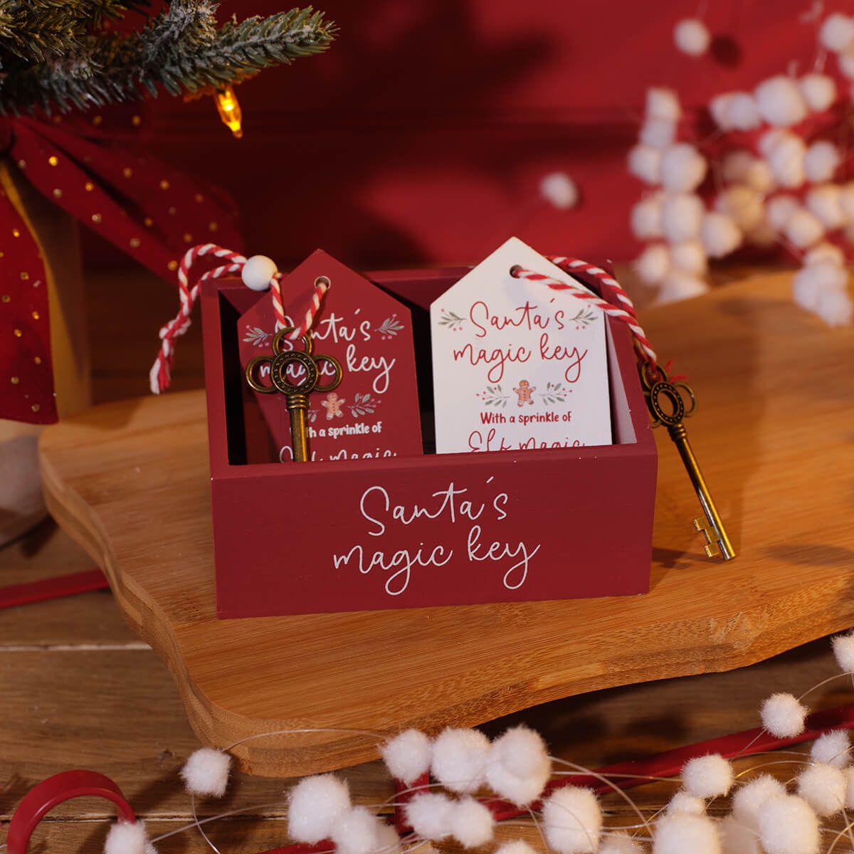 A red wooden box labeled 'Santa's magic key' on a wooden surface, with two hanging tags and decorative keys in a festive setting.