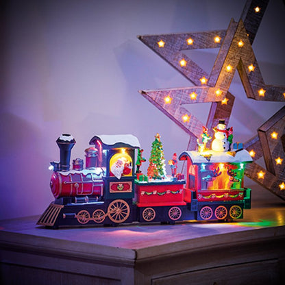 A festive Christmas-themed toy train set on a tabletop, featuring a red and black locomotive with Santa visible in the window, a small lit Christmas tree car, and a snowman figure on the rear car. The scene is styled with warm star-shaped marquee lights in the background.
