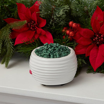 A white ribbed ceramic candle holder filled with green wax melts, styled on a white mantel with red poinsettias and green pine branches in the background.