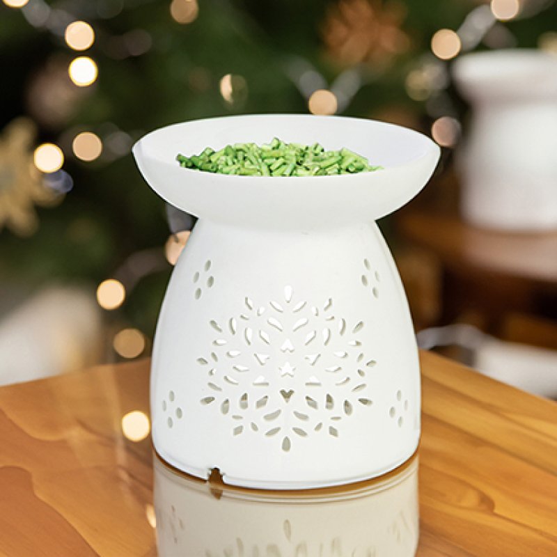 A white ceramic wax warmer with a snowflake cutout pattern sits on a wooden table, filled with green wax flakes, with a blurred Christmas tree in the background.