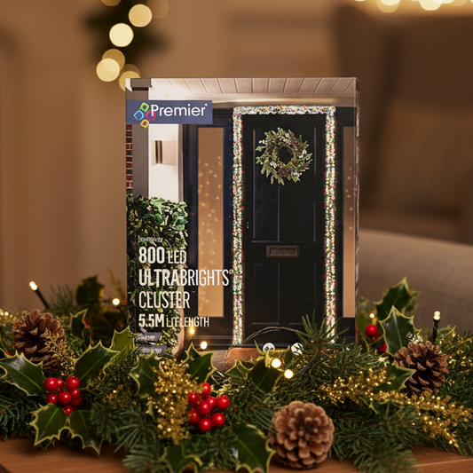 Premier 800 LED Ultrabrights cluster light in its clear plastic packaging, displayed on a festive Christmas garland with pinecones, holly, and red berries against a warm, blurred background.