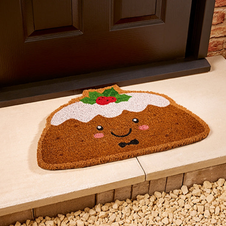 A brown coir doormat shaped like a smiling pudding with white frosting, green holly leaves, and a red berry topper, placed on a stone step in front of a dark door.