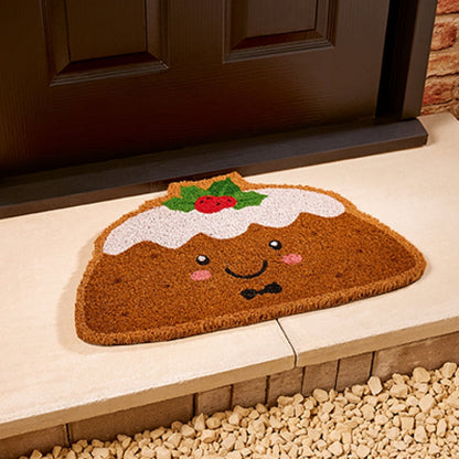 A brown coir doormat shaped like a smiling pudding with white frosting, green holly leaves, and a red berry topper, placed on a stone step in front of a dark door.