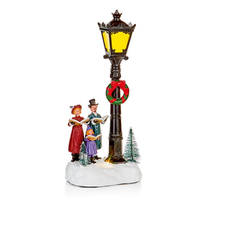 A festive Christmas village decorative lamp post figurine featuring two singing people and a child, a green wreath, and small pine trees on a snowy base, set against a plain white background.