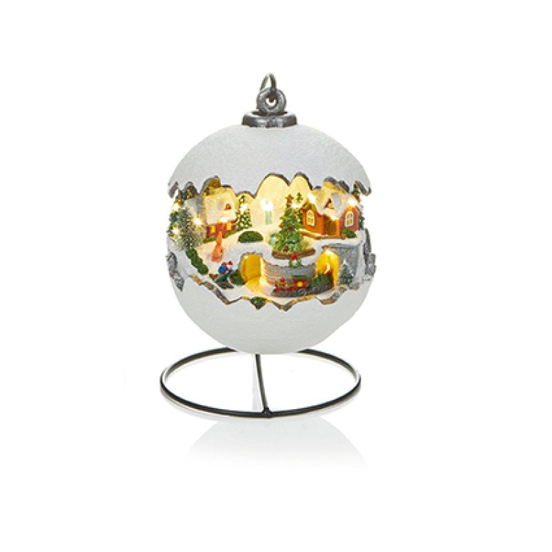 A white Christmas ornament shaped like an eggshell with a lit miniature village scene inside, displayed on a black metal stand against a white background.
