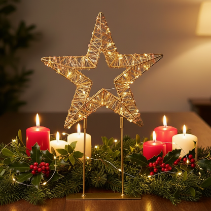 A glowing wire star decoration on a gold stand, lit with red and warm white fairy lights, displayed on a tabletop with green garland, pinecones, and red berries in the background.