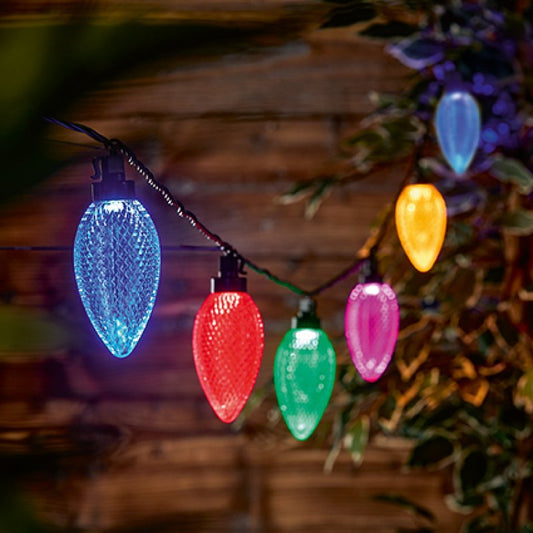 Multicolor Christmas string lights with textured teardrop-shaped bulbs in blue, red, green, pink, and yellow, hanging outdoors against a dark wooden fence and foliage.