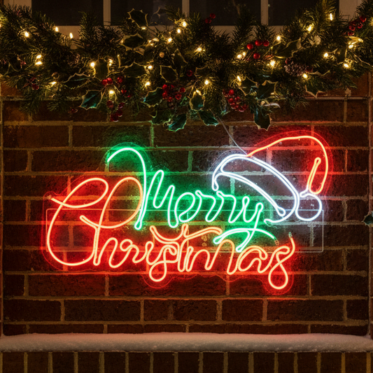 A glowing acrylic Christmas sign mounted on a brick wall, featuring the words “Merry Christmas” in red and green script with a white Santa hat. Festive green garland with warm white lights and red berries decorates the top of the brick wall above the sign.