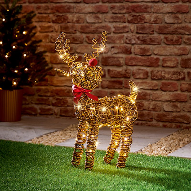 A golden wire reindeer yard decoration with warm white string lights and a red bow, standing on green grass in front of a brick wall.