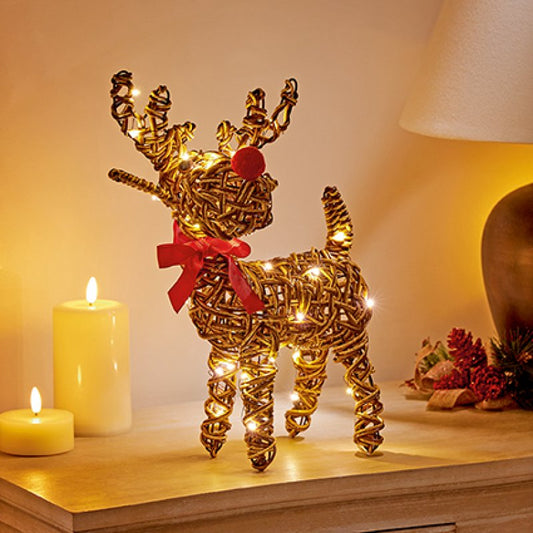 A golden wire reindeer Christmas decoration with spiral legs and antlers, lit with warm white fairy lights, wearing a red ribbon and red nose, displayed on a wooden table next to lit candles.