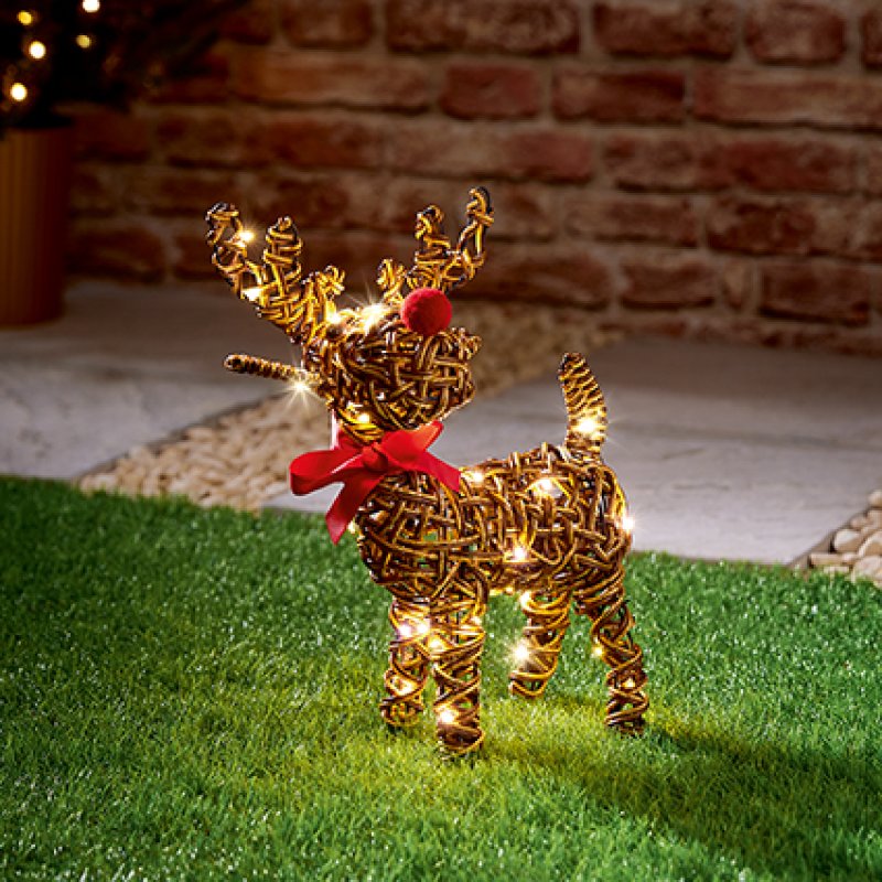 A golden wire reindeer yard light with warm white LED lights and a red pom-pom nose, standing on artificial grass in front of a brick wall.