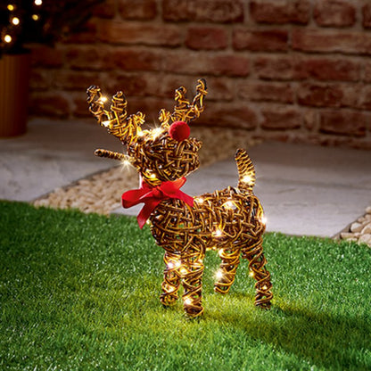 A golden wire reindeer yard light with warm white LED lights and a red pom-pom nose, standing on artificial grass in front of a brick wall.