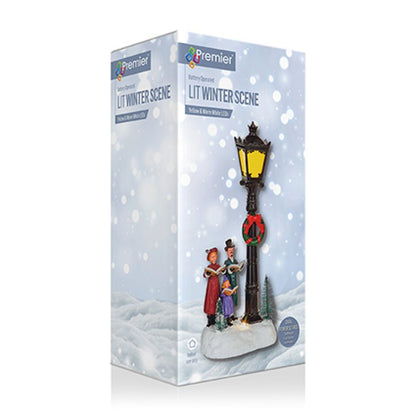 Premier Lighting 'Lit Winter Scene' decorative lamp post set in its retail packaging, featuring a black lamp post with a glowing yellow lantern, a red wreath, and two small holiday figures under falling snow graphics.
