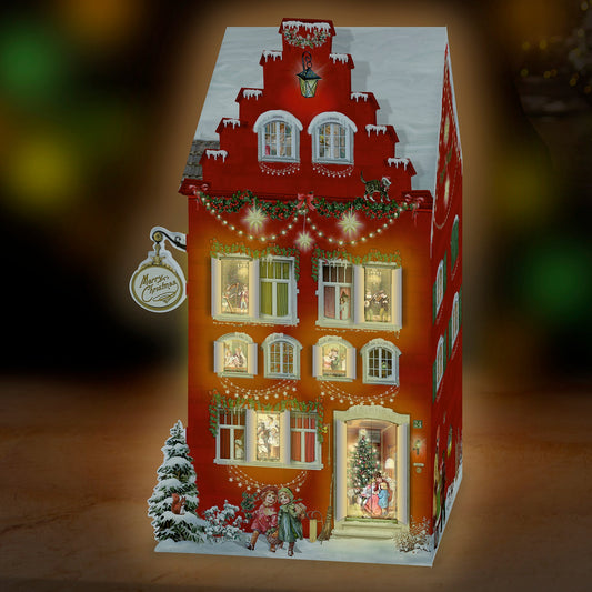 A decorative illuminated Christmas 3D house-shaped advent with a red façade, snow-dusted roof and trim, warm glowing windows, and small holiday figures and trees at the base, shown against a dark blurred background.