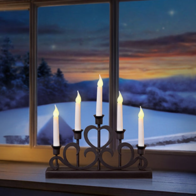 A five-light black metal candle holder with a heart-shaped center detail sits on a windowsill, casting a warm glow against a window showing a snowy winter landscape at dusk.