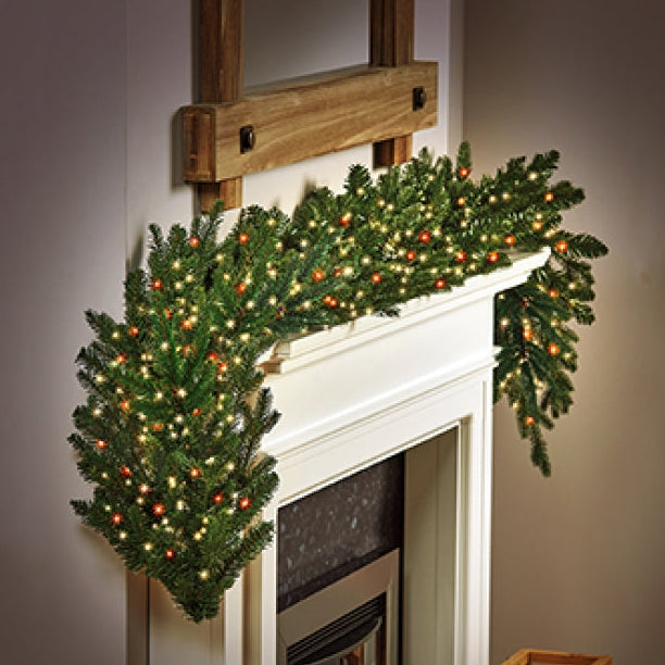 A green holiday garland with warm white  and red string lights draped over a white fireplace mantel in a cosy indoor setting.