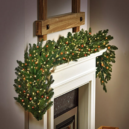 A green holiday garland with warm white  and red string lights draped over a white fireplace mantel in a cosy indoor setting.
