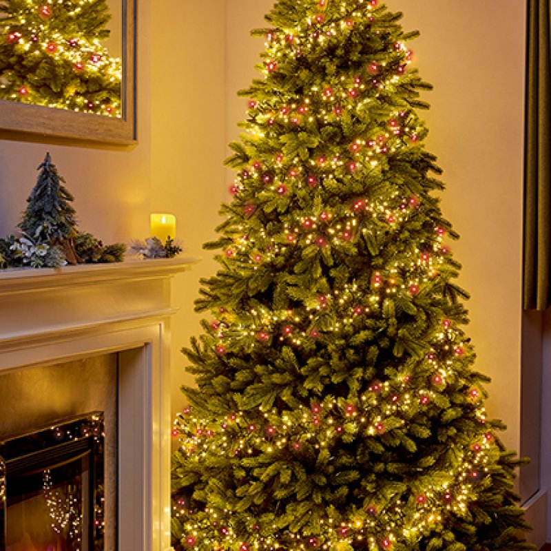 A tall artificial Christmas tree decorated with red and warm white string lights and small red ornaments, standing beside a fireplace in a cosy indoor setting.