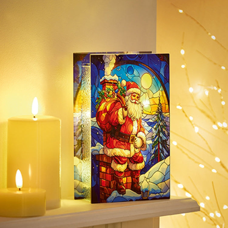 A rectangular decorative plaque or sign featuring a stained-glass-style illustration of Santa Claus in a chimney scene, displayed on a shelf beside lit candles and warm string lights.