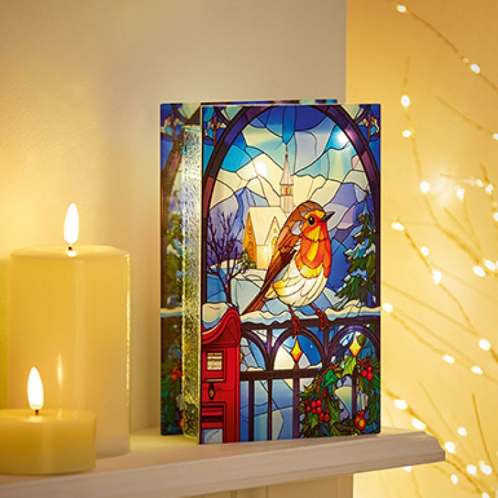 A rectangular stained-glass-style art panel featuring a robin bird in front of a church spire and fence, displayed on a shelf beside lit candles and warm string lights.