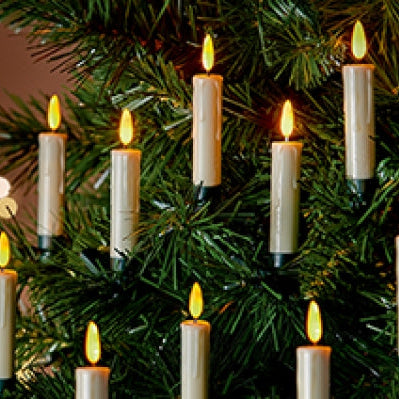 A close-up of a Christmas tree decorated with multiple lit white pillar candles with glowing yellow flames, set against a softly blurred warm indoor background with bokeh lights.