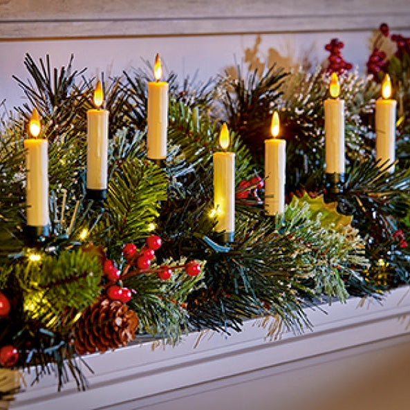 A festive Christmas garland draped across a white fireplace mantel, featuring green pine branches, red berries, pinecones, and multiple lit cream pillar candles.