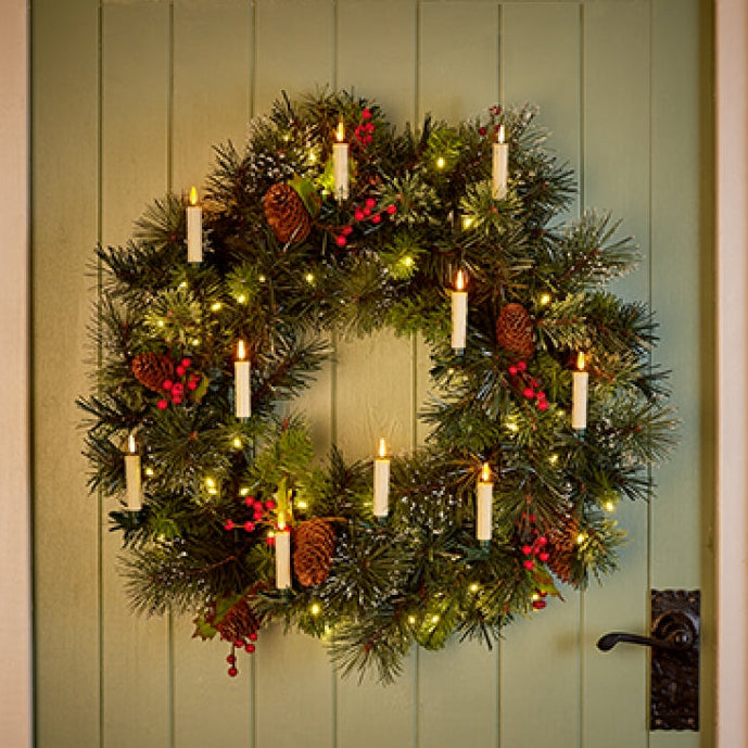 A festive green wreath with lit white pillar candles, pinecones, and red berries hanging on a light sage green paneled door.