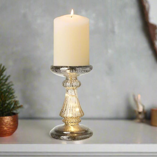 A lit gold glass candle holder with a decorative base, holding a candle, placed on a white surface with a tree in the background.