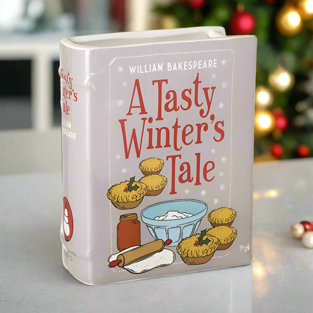 Ceramic container with 'A Tasty Winter's Tale' design featuring pies and cooking elements, set against a blurred festive background.