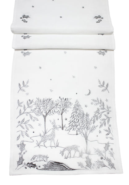 A white fabric table runner, featuring a delicate silver embroidered woodland scene with trees, deer, stars, and a crescent moon.