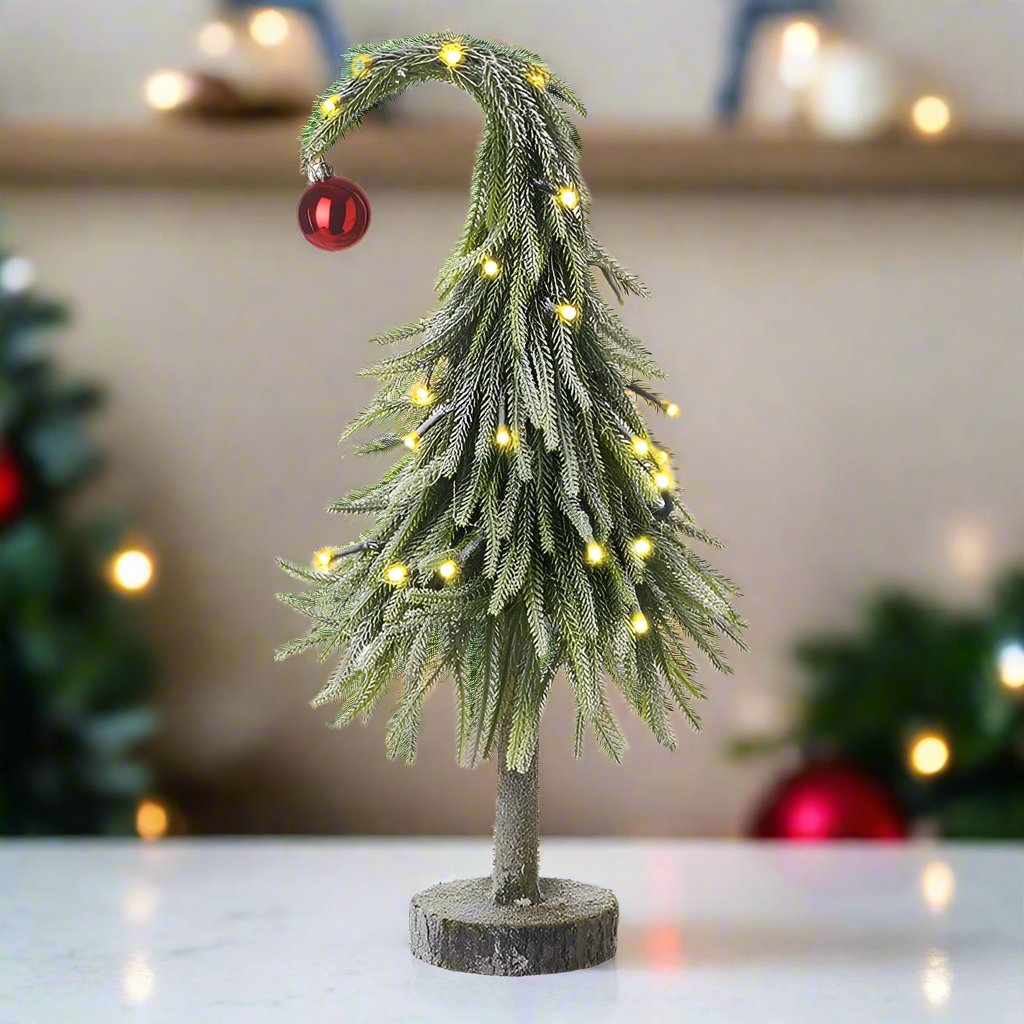 A bending artificial Christmas tree with warm white LED lights, placed on a wooden stand, with a red ornament at the top.