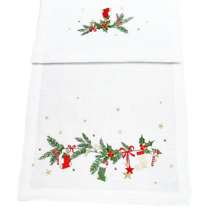 A white table runner with embroidered Christmas garlands, including green holly and pine branches, red bows, red stockings, and small gold stars.