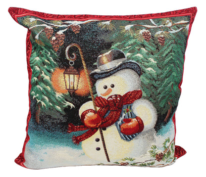 A square decorative throw pillow featuring a festive needlepoint-style winter scene with a snowman wearing a red scarf and hat, surrounded by pine trees, snow, and a glowing lantern. The pillow has a red piped border and is shown against a plain white background.