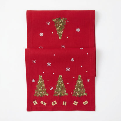 A red Christmas table runner featuring three Christmas trees with small gold gift boxes and white sequin snowflakes on a plain white background.