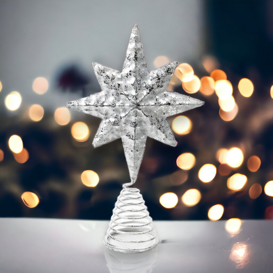 A rustic silver metal star-shaped Christmas tree topper with a pressed design and embossed textures, placed on a white surface with blurred festive lights in the background.