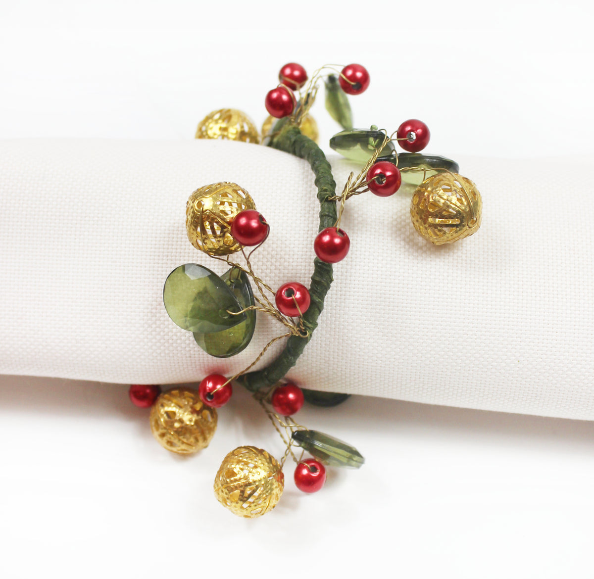 A festive napkin ring with gold textured bells, red beads, green leaves, and a dark green vine, displayed on a white napkin.