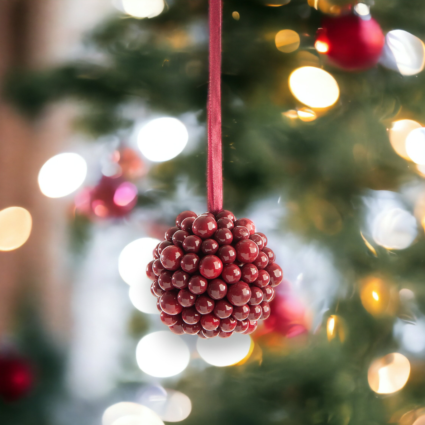 A close-up image of a burgundy berry cluster ball Christmas decoration with a blurred Christmas tree and lights in the background.