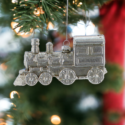 A silver hanging decoration in the shape of a train, hanging from a Christmas tree with lights and red decorations in the background.