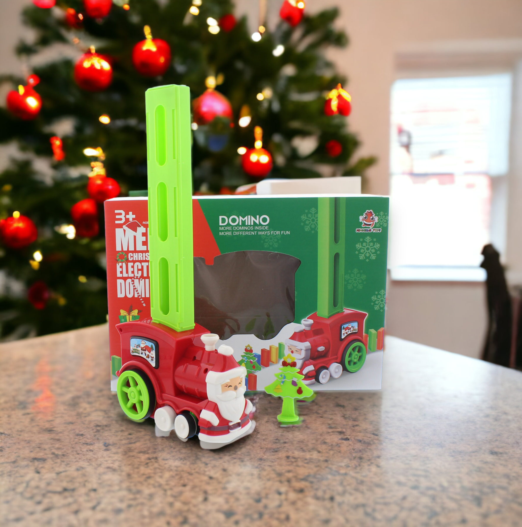 A battery-operated electronic domino train toy set on a table, with red and green domino pieces, and a Christmas tree in the background.