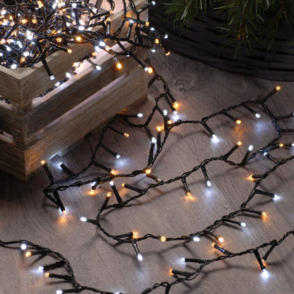 A set of festive Christmas tree lights with a mix of white and warm white bulbs, arranged on a wooden surface, indoors, with a Christmas tree in the background.