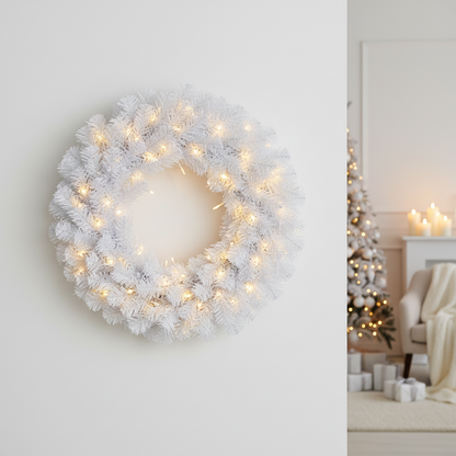 A white artificial Christmas wreath with warm white string lights, mounted on a light gray wall. In the softly blurred background on the right, a decorated Christmas tree and cozy living room setting are visible.