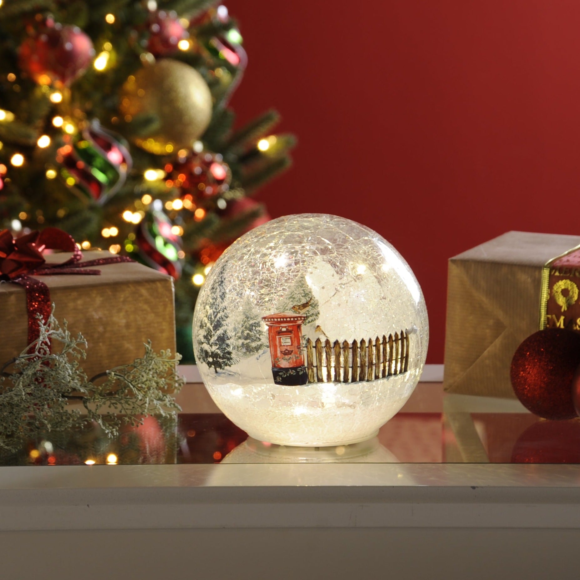 A lit crackle effect Christmas ball with a winter scene post-box, placed on a table with Christmas decorations in the background.