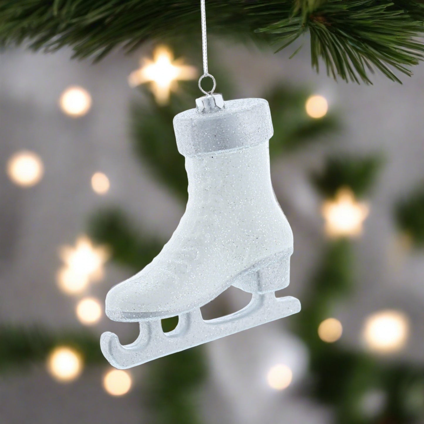 A white glittery hanging Christmas decoration shaped like a skate, with a silver hanging wire against a blurred Christmas tree background.