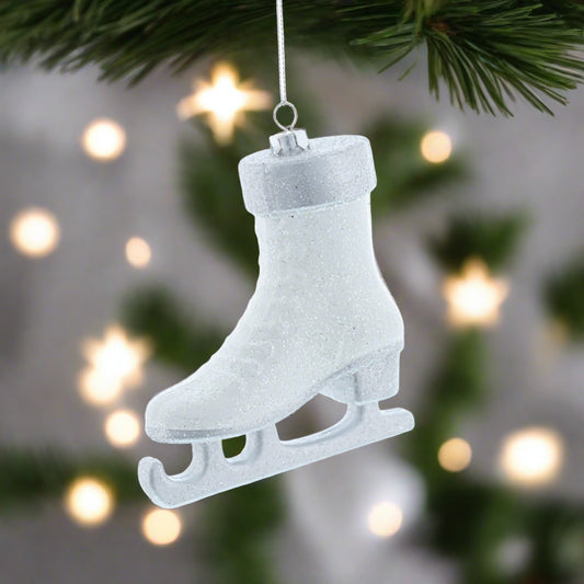 A white glittery hanging Christmas decoration shaped like a skate, with a silver hanging wire against a blurred Christmas tree background.
