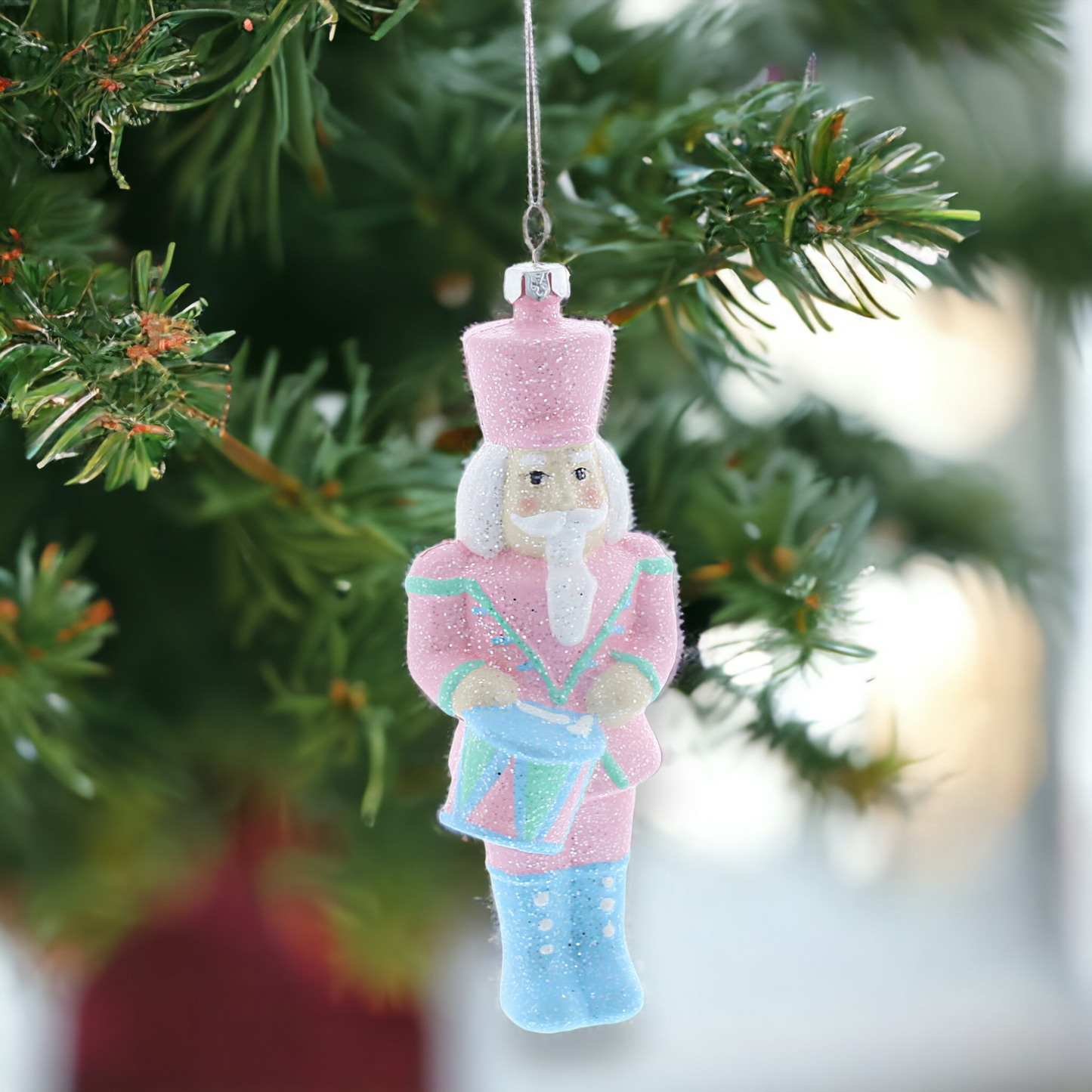 A pastel-colored nutcracker hanging decoration with glitter, placed on a Christmas tree with a blurred green background.