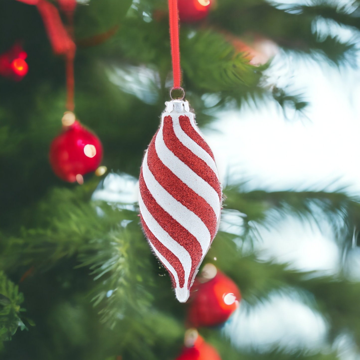 A red and white striped glitter hanging decoration resembling a candy, placed on a Christmas tree with red ornaments and green leaves.